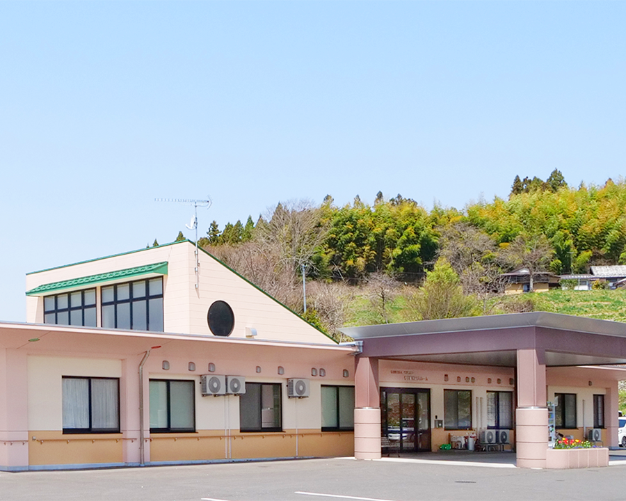 介護施設の建物外観。ピンク色の壁と緑色の屋根、背景に緑の山々が見える施設全景