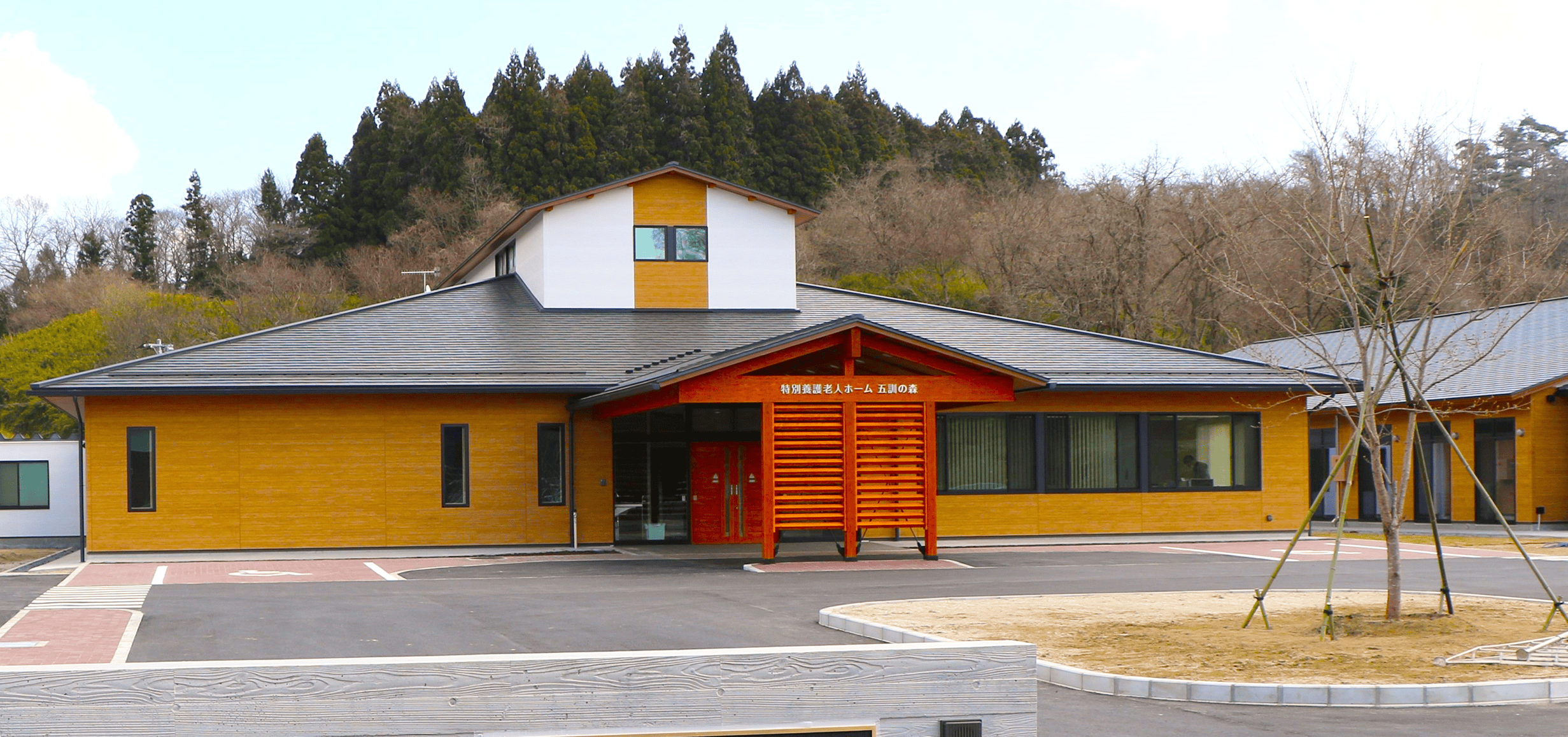 黄色と赤のモダン外観が特徴的な介護施設。周囲を緑に囲まれた立地。